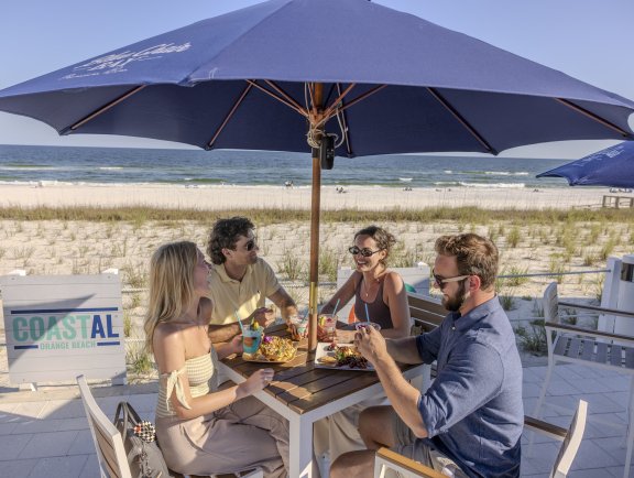 Friends dining under an umbrella at Coastal beachfront restaurant in Orange Beach