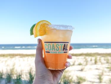 Hand holding a bright orange cocktail in front of the beach at Coastal Orange Beach restaurant