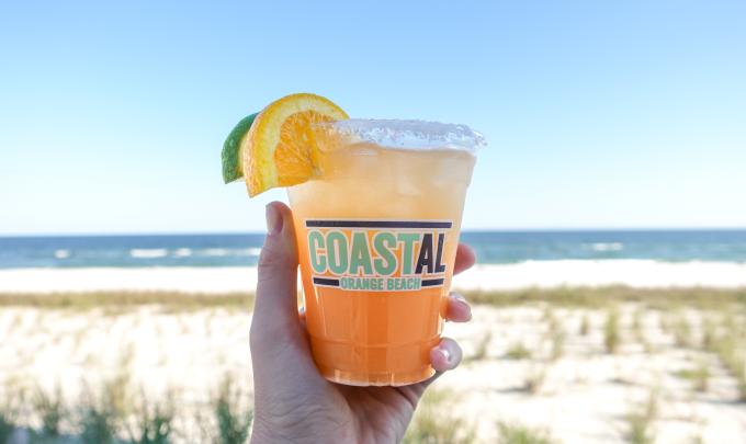 Hand holding a bright orange cocktail in front of the beach at Coastal Orange Beach restaurant