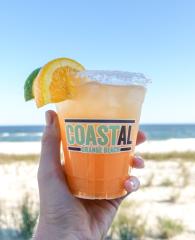 Hand holding a bright orange cocktail in front of the beach at Coastal Orange Beach restaurant