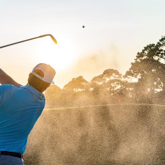 Golfer hitting a sand shot from a bunker at Kiva Dunes, with sand and dust exploding around the ball and club face.