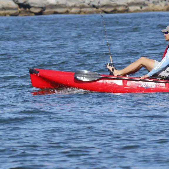 Man wearing sunglasses and a hat kayak fishing in a red sit-on-top fishing kayak with multiple rods and a cooler on Alabama's Beaches.