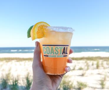 Hand holding a bright orange cocktail in front of the beach at Coastal Orange Beach restaurant