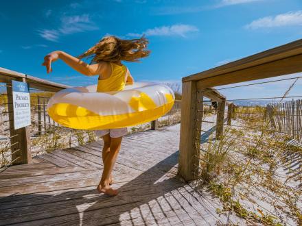 Young girl running on a boardwalk with a pool float toward the beach