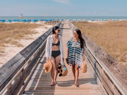 Two women walking down a boardwalk at the beach with straw hats and beach bags