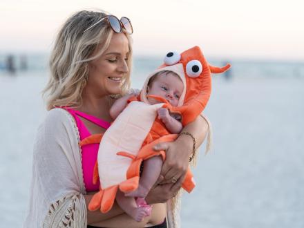 A woman holding a baby in a shrimp costume on the beach during the National Shrimp Festival in Gulf Shores