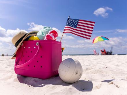 pink beach bag filled with beach gear and an American flag sitting next to a volleyball on the sand