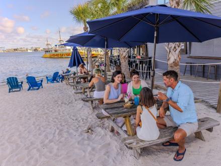 People dining at tables in the sand by the water at Avenue Pub casual outdoor restaurant in Orange Beach