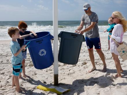 family putting items in garbage and recycling bins while on the beach in Gulf Shores
