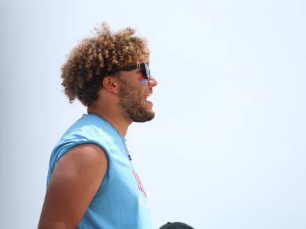 Side profile of an excited male fan with curly hair and sunglasses, cheering at the NCAA Beach Volleyball Championship.
