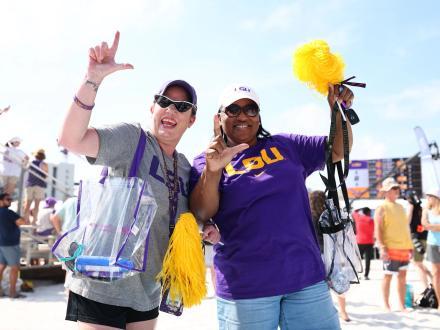 Two excited LSU volleyball fans cheering with pom-poms and making the 'L' sign at the NCAA Beach Volleyball Championship in Gulf Shores, Alabama.