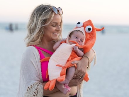 A woman holding a baby in a shrimp costume on the beach during the National Shrimp Festival in Gulf Shores